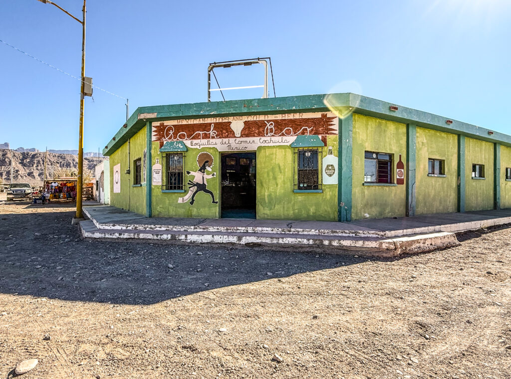 Exterior view of Boquillas del Carmen restaurant with green walls and decorative signage, set against a clear blue sky in a rural landscape.