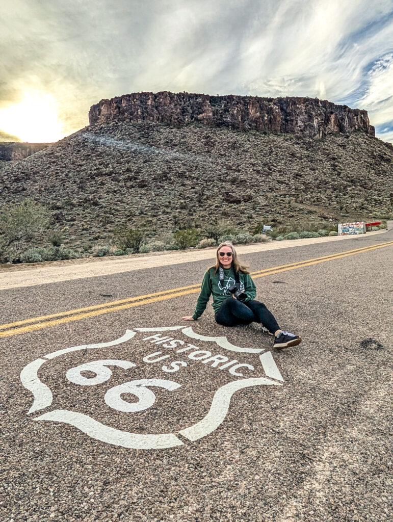 Traveler sitting beside a painted Historic Route 66 sign on the road, with rugged mountains in the background under a cloudy sky.