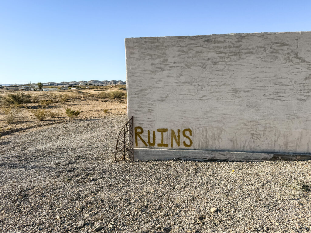 A close-up of a weathered wall with the word 'RUINS' painted in yellow, set against a backdrop of a desert landscape with sparse vegetation and mountains in the distance.